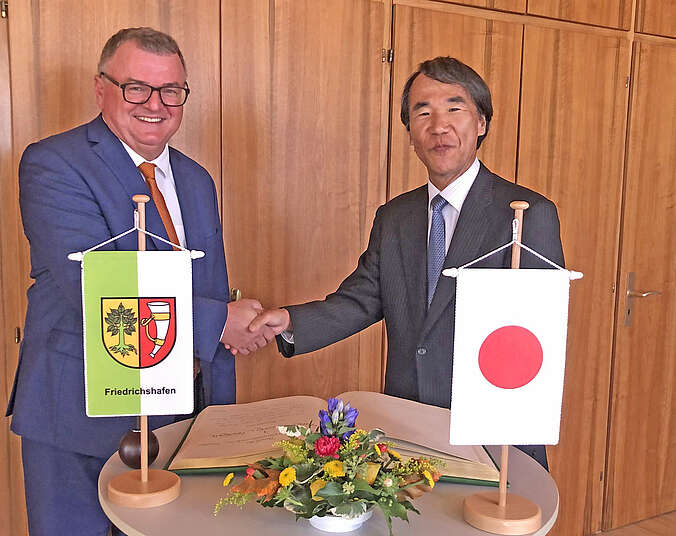 OB Brand und der Generalkonsul aus Japan geben sich die Hände. Davor stehen zwei Wimpel von Friedrichshafen und Japan