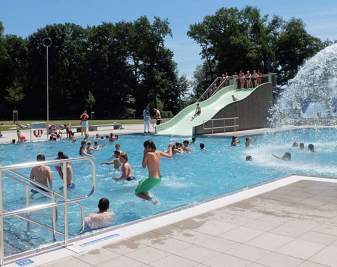 Becken mit blauem Wasser und vielen Kindern und Erwachsenen im Wasser. Im Hintergrund strudelt ein Wasserpilz.