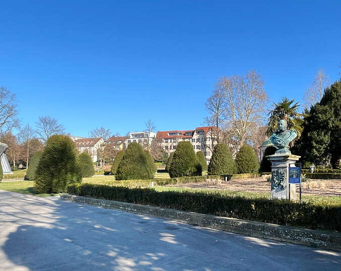 Uferpark mit Pflanzbeeten, Büste, im Hintergrund Musikmuschel, Bäume und Häuser