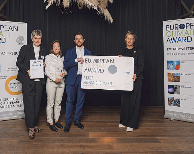 Erster Bürgermeister Fabian Müller und Kira Urban nehmen große Tafel, Award und Urkunde auf Bühne von zwei Frauen entgegen.